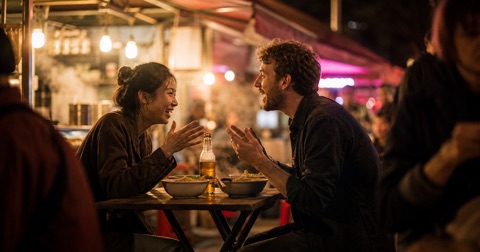 Two friends talking over bowls of noodles at a night-time Chinese street-food stall.